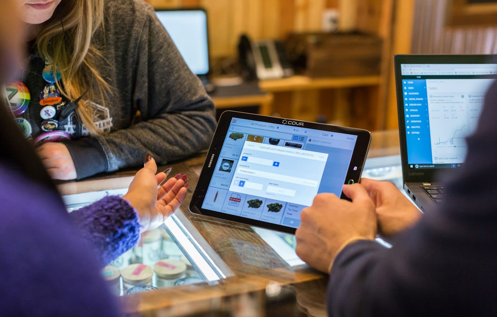 At a shop counter, a staff member adds a product on a tablet point of sale while the laptop beside them shows a sales dashboard